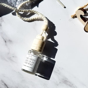 May include: A white string with a small glass bottle hanging from it. The bottle is clear and has a white label that says "Nordic Sentiments Autumn Collection Northern Lights in Norway". The bottle is sitting on a white marble surface.