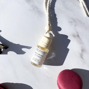 May include: A white string car air freshener with a wooden bead and a clear glass bottle with a white label that reads "Even Continous Collection Macarons in Paris 10 ml / 0.33 fl oz Made in Canada". The bottle is surrounded by macarons in various colors, including pink, beige, and yellow.