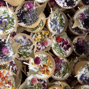 May include: A collection of small, round, white candles with dried flowers and herbs embedded in the wax. The candles are in paper cups with a natural brown color.