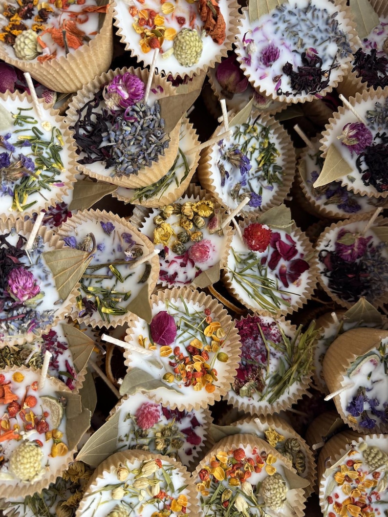 May include: A collection of small, round, white candles with dried flowers and herbs embedded in the wax. The candles are in paper cups with a natural brown color.