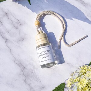 May include: A small, clear glass bottle with a beige cap and a white label that reads "Nantucket Hydrangea House." The bottle hangs from a cream-colored rope with a wooden bead. The background features white and gray marble and hydrangea flowers.