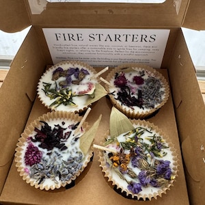 May include: Four fire starters in a cardboard box. The fire starters are made of natural waxes like soy, coconut, or beeswax. They are decorated with dried flowers and herbs. The box has the text "FIRE STARTERS" printed on it.