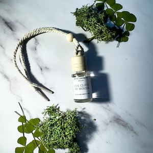 May include: A white string car air freshener with a clear glass bottle containing a fragrance called "The Cliffs of Moher" by Seven Continents Ireland. The bottle is on a white marble surface with green moss and clover leaves.