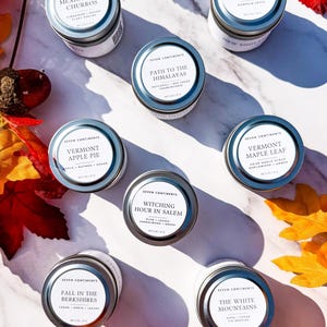 May include: An overhead shot of several small, round metal tins with white labels. The labels feature text like "Mexico City Churros" and "Vermont Apple Pie." Autumn leaves and berries surround the tins, creating a seasonal display.
