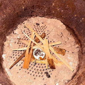 May include: A close-up view of a metal smoker with a perforated grate and a layer of ash.  A small pile of kindling wood is arranged on top of the grate.