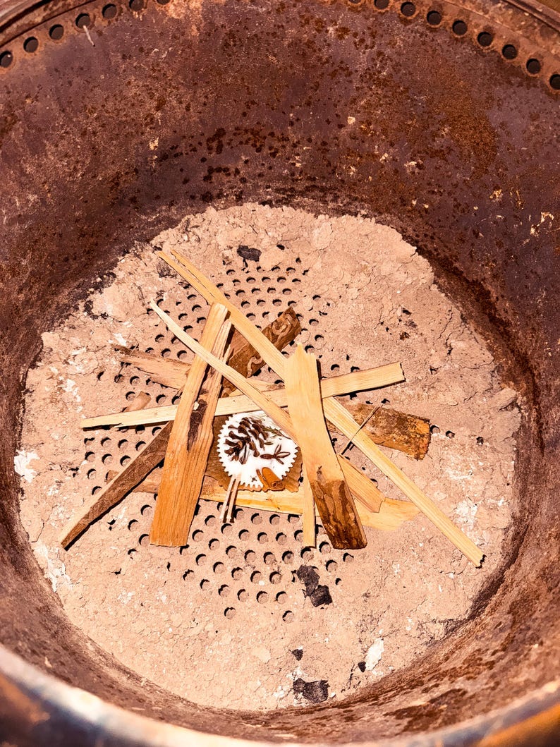 May include: A close-up view of a metal smoker with a perforated grate and a layer of ash.  A small pile of kindling wood is arranged on top of the grate.