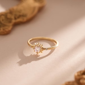 Könnte beinhalten: Ein goldener Ring mit einem klaren Edelstein in der Mitte, umgeben von kleineren Edelsteinen. Der Ring hat ein zartes, strukturiertes Band.