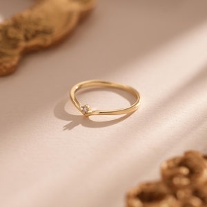 May include: A delicate gold ring with a small, sparkling gemstone at the centre. The ring has a V-shaped design and a thin band. The background features a soft, neutral colour palette with a blurred gold decorative element.