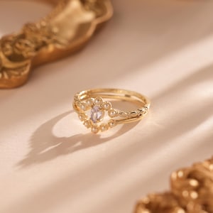 Può includere: Un anello dorato con una pietra centrale ovale, viola chiaro, circondata da piccole perle. L'anello ha un design delicato e ornato. È appoggiato su una superficie rosa chiaro, con una cornice decorativa dorata sullo sfondo.