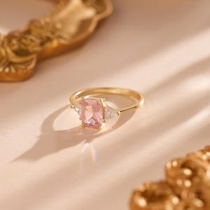 Könnte beinhalten: Ein goldener Ring mit einem rechteckigen rosa Edelstein in der Mitte und zwei kleineren dreieckigen weißen Edelsteinen auf beiden Seiten. Der Ring befindet sich vor einem weichen rosa Hintergrund mit goldenen Dekorationselementen.