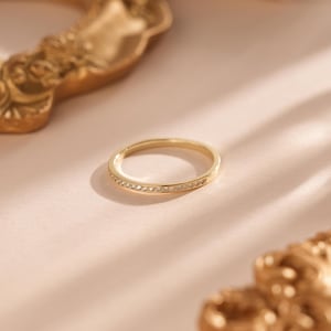 Puede incluir: Un anillo de banda de oro con pequeñas piedras preciosas transparentes engastadas en la parte superior. El anillo está sobre una superficie rosa claro, con un marco decorativo dorado borroso en el fondo. Es una joya delicada.