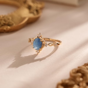 Könnte beinhalten: Ein goldfarbener Ring mit einem ovalen, hellblauen Edelstein in der Mitte. Der Ring hat eine dekorative Fassung mit kleinen, klaren Akzentsteinen. Das Band hat ein aufwendiges Design. Der Ring ist vor einem weichen, neutralen Hintergrund platziert.