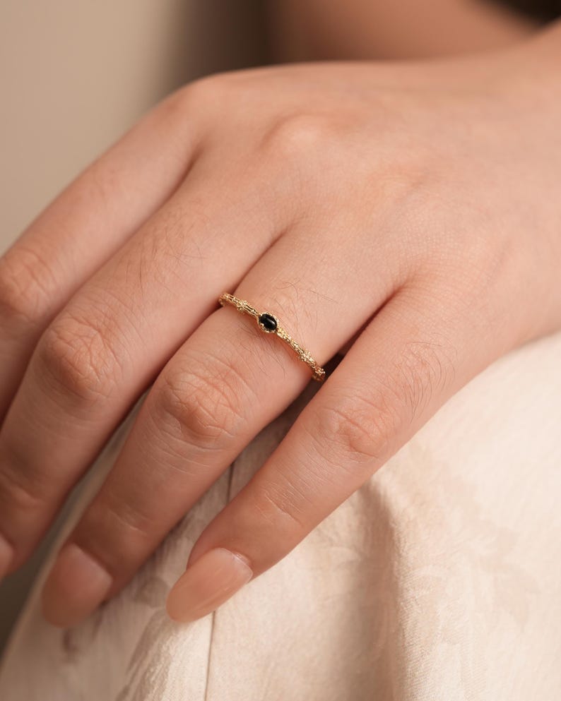 Puede incluir: Un delicado anillo de oro con una piedra preciosa negra engastada en el centro. El anillo est&aacute; hecho de una banda texturizada.