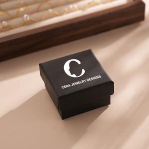 Puede incluir: Una caja de joyer&iacute;a negra con un logotipo blanco que dice "CERA JEWELRY DESIGNS" y una silueta estilizada de una cabeza de persona con una "C" dentro.