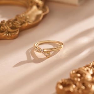 Puede incluir: Un anillo dorado con un diseño de lazo, adornado con pequeñas piedras brillantes. El anillo se encuentra sobre un fondo suave y neutro, con un marco dorado decorativo en el fondo. El delicado diseño del anillo lo convierte en un accesorio perfecto.