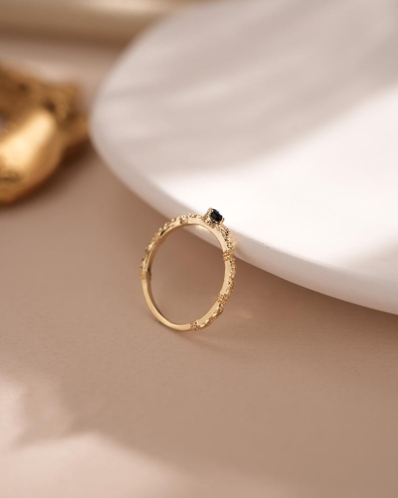 Puede incluir: Un anillo de oro con una banda delicada y texturizada y una peque&ntilde;a piedra preciosa negra en el centro.