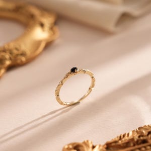 Puede incluir: Un delicado anillo de oro con una peque&ntilde;a piedra preciosa negra engastada en el centro. El anillo tiene una banda texturizada con un dise&ntilde;o floral.