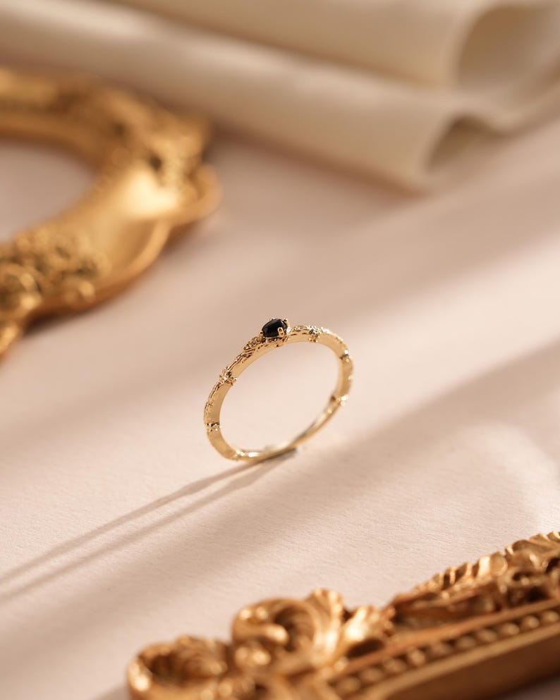 Puede incluir: Un delicado anillo de oro con una peque&ntilde;a piedra preciosa negra engastada en el centro. El anillo tiene una banda texturizada con un dise&ntilde;o floral.