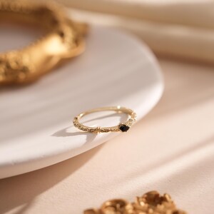 Puede incluir: Un anillo de oro con una banda delicada y texturizada y una peque&ntilde;a piedra preciosa negra. El anillo est&aacute; sobre una superficie blanca.