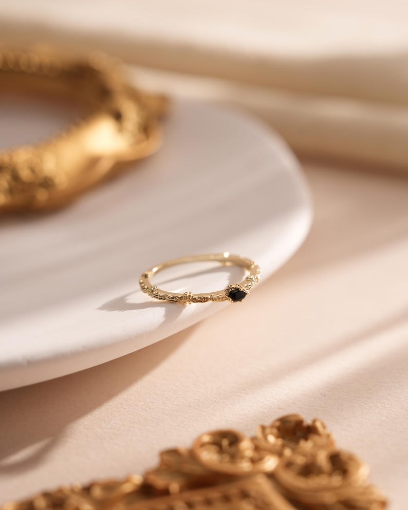 Puede incluir: Un anillo de oro con una banda delicada y texturizada y una peque&ntilde;a piedra preciosa negra. El anillo est&aacute; sobre una superficie blanca.