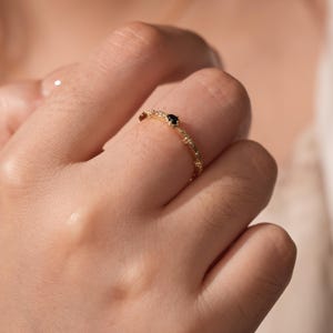 Puede incluir: Un delicado anillo de oro con una peque&ntilde;a piedra preciosa negra engastada en el centro. El anillo tiene una banda texturizada con un dise&ntilde;o delicado.