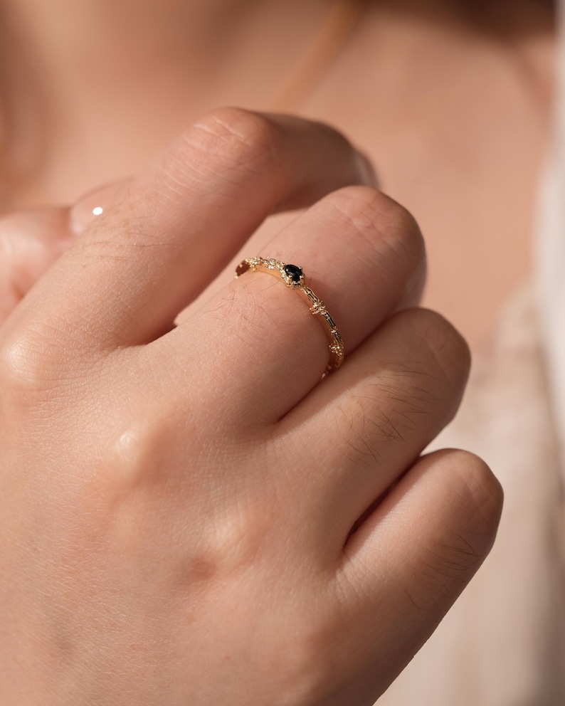 Puede incluir: Un delicado anillo de oro con una peque&ntilde;a piedra preciosa negra engastada en el centro. El anillo tiene una banda texturizada con un dise&ntilde;o delicado.