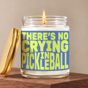 May include: A clear glass candle jar with a lit wick and a gold-colored metal lid resting beside it. The label features a blue background with the lime green text "THERE'S NO CRYING IN PICKLEBALL" printed in a bold, sans-serif, block-style font.