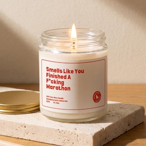 May include: A lit soy wax candle in a clear glass jar with a white label featuring red text that reads: Smells Like You Finished A F*cking Marathon. The label also includes the text 100% Soy Wax Candle, Handpoured in California, B201, 03. Zen.