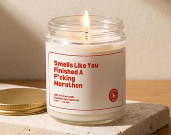 May include: A lit soy wax candle in a clear glass jar with a white label featuring red text that reads: Smells Like You Finished A F*cking Marathon. The label also includes the text 100% Soy Wax Candle, Handpoured in California, B201, 03. Zen.