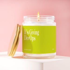 May include: A lit candle in a glass jar with a gold lid. The jar has a bright green label with the text "It's Giving DevOps" in white. The candle is on a white surface against a pink background.