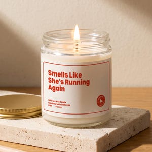 May include: A clear glass jar candle with a lit wick and white wax. The label features a red border and red text that reads: Smells Like She's Running Again, 100% Soy Wax Candle, Handpoured in California, B180, 03. Zen, and a small circular logo.