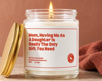 May include: A clear glass jar candle with a lit wick and a gold-colored metal lid. The white label features red text that reads: Mom, Having Me As A Daughter Is Really The Only Gift You Need, along with 100% Soy Wax Candle, Handpoured in California.