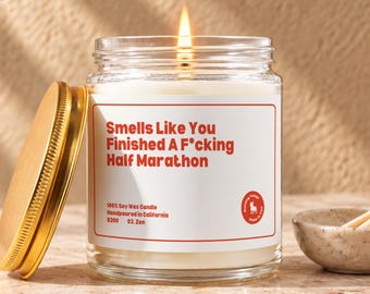 May include: A clear glass jar candle with a lit wick and a gold-colored metal lid. The white label features red text reading Smells Like You Finished A F*cking Half Marathon, 100% Soy Wax Candle, Handpoured in California, B200, 03. Zen, and a logo.
