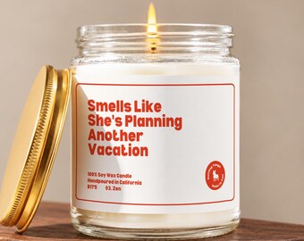 May include: A clear glass jar candle with a lit wick and a gold-colored metal lid. The white label features red text reading Smells Like She's Planning Another Vacation, 100% Soy Wax Candle, Handpoured in California, B175, 03. Zen, and a logo.