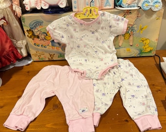 Puede incluir: Un conjunto de ropa de bebé con un body blanco y pantalones a juego. El body tiene un ribete rosa y está estampado con pequeñas imágenes repetidas. Los pantalones son de color rosa liso. El conjunto se muestra en una percha amarilla.