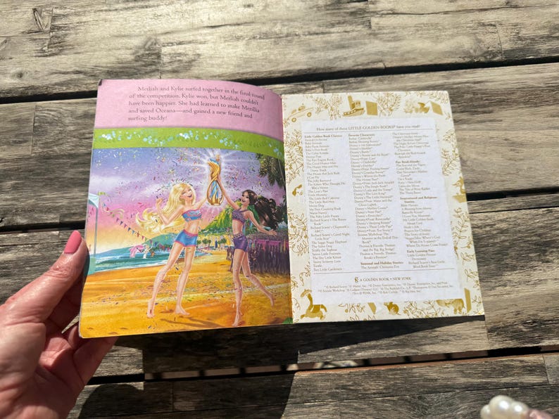 Pode incluir: Livro aberto com ilustra&ccedil;&otilde;es coloridas. Uma p&aacute;gina mostra duas mulheres numa praia, segurando um trof&eacute;u. A outra p&aacute;gina lista t&iacute;tulos. O livro tem uma borda rosa e o texto "Little Golden Book".