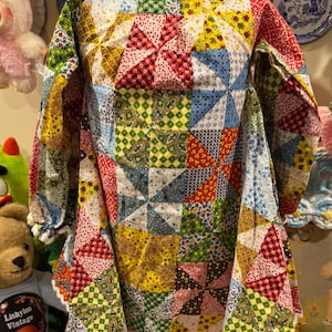 May include: A vintage patchwork dress with long sleeves, featuring a colorful pinwheel design. The dress is made of various patterned fabrics in red, yellow, blue, and green, with white lace trim around the collar and cuffs. The dress is displayed on a mannequin.