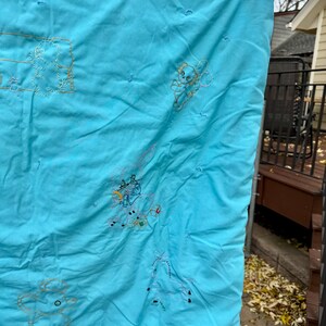 May include: A light blue quilt with embroidered cartoon characters. The quilt is hanging outside on a clothesline. The embroidery is in shades of gold and white. The quilt has a quilted pattern.