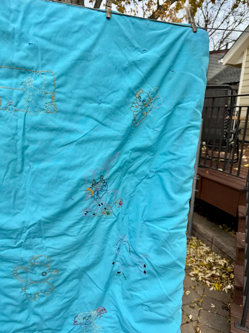 May include: A light blue quilt with embroidered cartoon characters. The quilt is hanging outside on a clothesline. The embroidery is in shades of gold and white. The quilt has a quilted pattern.