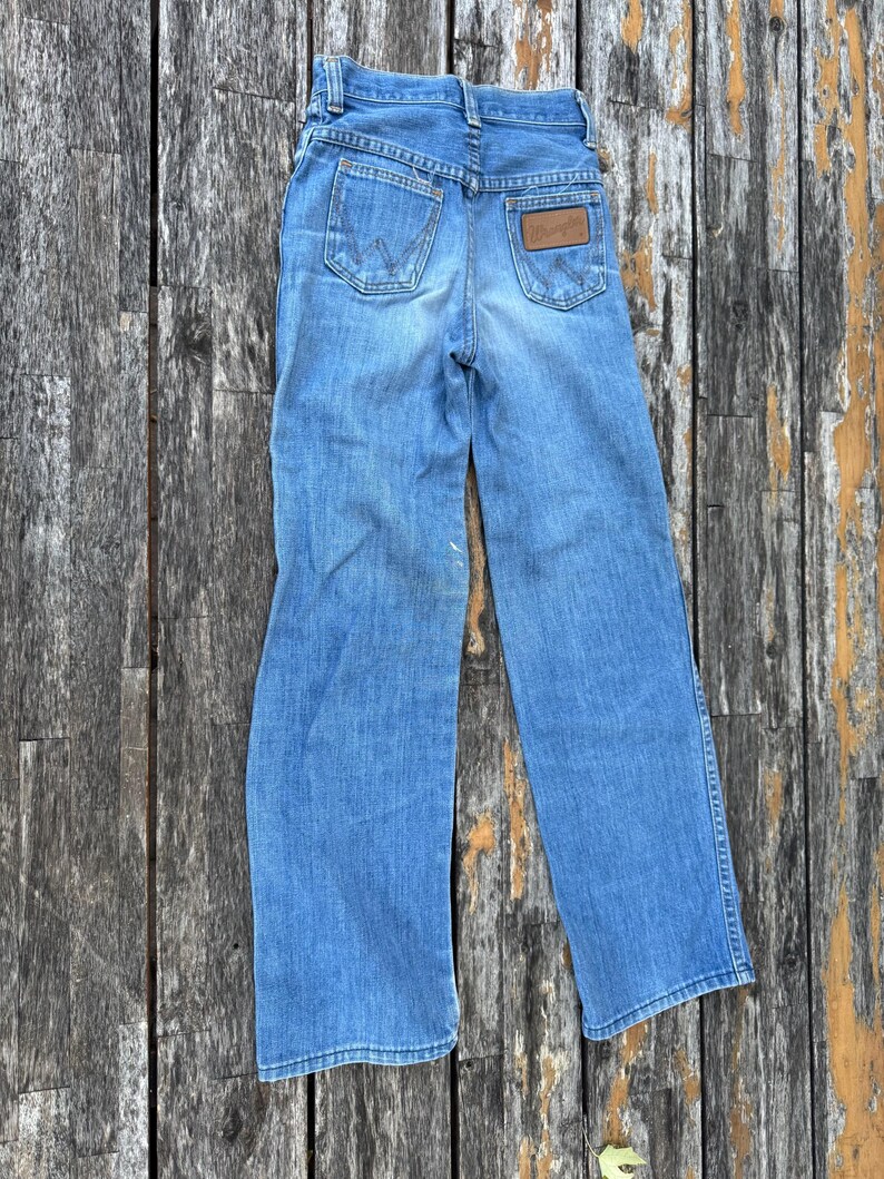 Puede incluir: Vaqueros de mezclilla azul claro con un dise&ntilde;o cl&aacute;sico de cinco bolsillos. La parte trasera presenta dos bolsillos y un parche de cuero marr&oacute;n con el logotipo "Wrangler". Los vaqueros est&aacute;n sobre una superficie de madera desgastada.