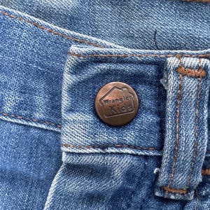 Puede incluir: Primer plano de unos vaqueros azules con un bot&oacute;n de color cobre. El bot&oacute;n tiene el logotipo de Wrangler Kids en relieve. Los vaqueros tienen costuras marrones y un dise&ntilde;o cl&aacute;sico de cinco bolsillos.