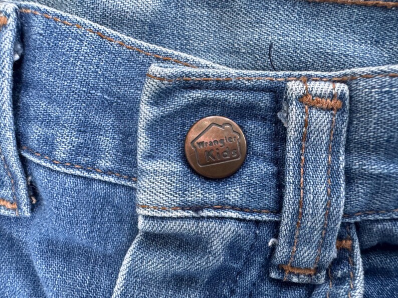 Puede incluir: Primer plano de unos vaqueros azules con un bot&oacute;n de color cobre. El bot&oacute;n tiene el logotipo de Wrangler Kids en relieve. Los vaqueros tienen costuras marrones y un dise&ntilde;o cl&aacute;sico de cinco bolsillos.