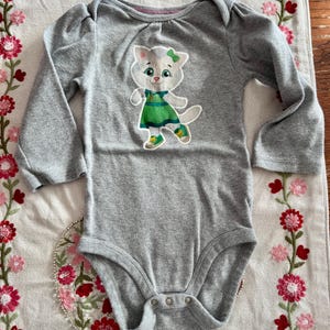 May include: Gray long-sleeved baby bodysuit with a cartoon cat graphic. The cat wears a green dress and shoes, with a green bow. The bodysuit has a round neckline and snap closures at the bottom.