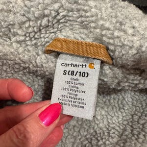 Puede incluir: Primer plano de una etiqueta Carhartt, talla S (8/10), con detalles de la composici&oacute;n de la tela. La etiqueta est&aacute; adherida a una tela gris texturizada. La etiqueta dice "Shell: 100% Cotton, Lining: 100% Polyester, Filling: 100% Polyester, Exclusive of trims, Made in Vietnam."