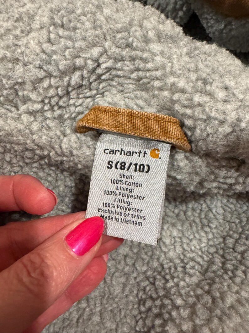 Puede incluir: Primer plano de una etiqueta Carhartt, talla S (8/10), con detalles de la composici&oacute;n de la tela. La etiqueta est&aacute; adherida a una tela gris texturizada. La etiqueta dice "Shell: 100% Cotton, Lining: 100% Polyester, Filling: 100% Polyester, Exclusive of trims, Made in Vietnam."