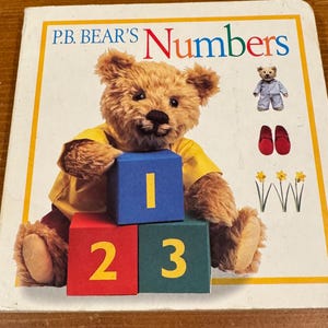 May include: A children's book titled "P.B. Bear's Numbers" featuring a plush bear holding numbered blocks. The cover includes illustrations of a small bear, red shoes, and daffodils. The numbers 1, 2, and 3 are displayed.