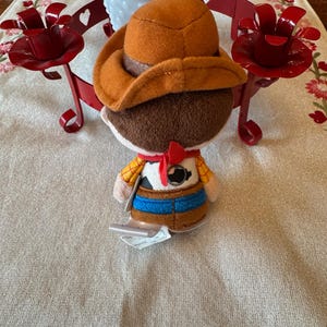 May include: A plush toy cowboy character, wearing a brown hat, a yellow shirt with a cowhide vest, a blue belt, and a red neckerchief. The toy has a tag attached. The background includes red candle holders and a white vase.