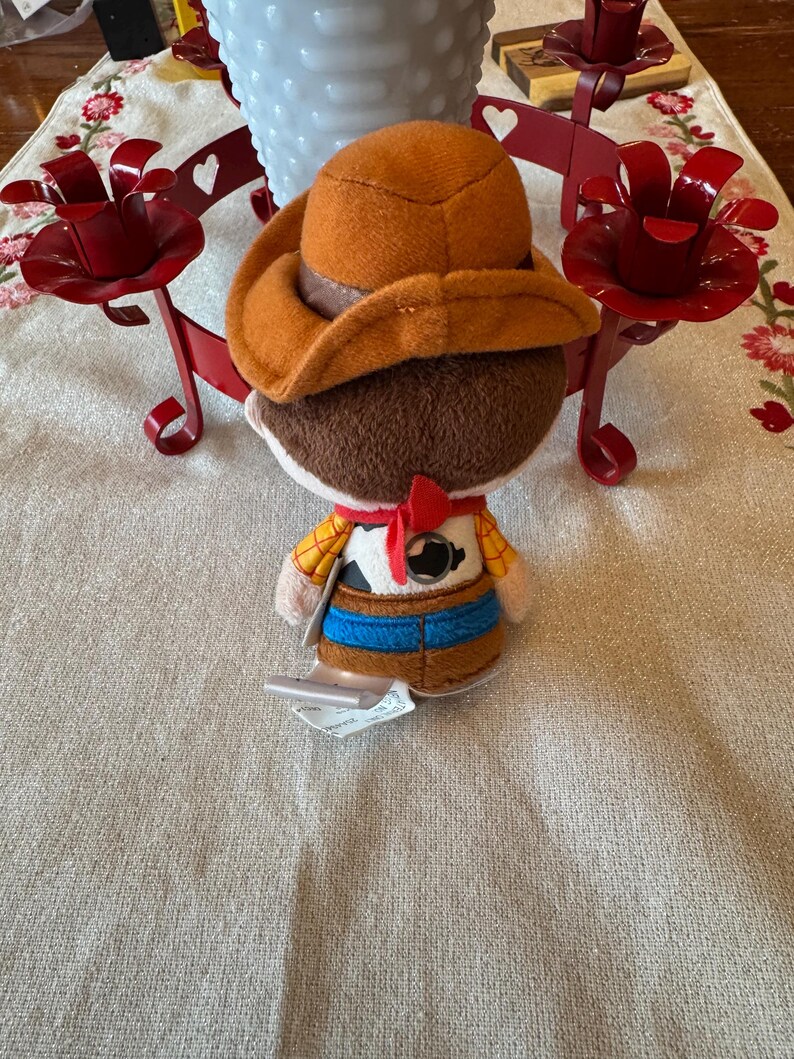 May include: A plush toy cowboy character, wearing a brown hat, a yellow shirt with a cowhide vest, a blue belt, and a red neckerchief. The toy has a tag attached. The background includes red candle holders and a white vase.