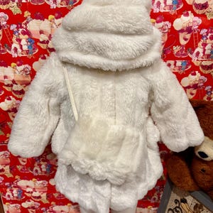May include: A fluffy white hooded jacket and skirt set. The jacket has long sleeves, a front pocket, and a drawstring. The skirt has a scalloped hem. The outfit is made of a soft, plush material, perfect for a child's winter wardrobe.