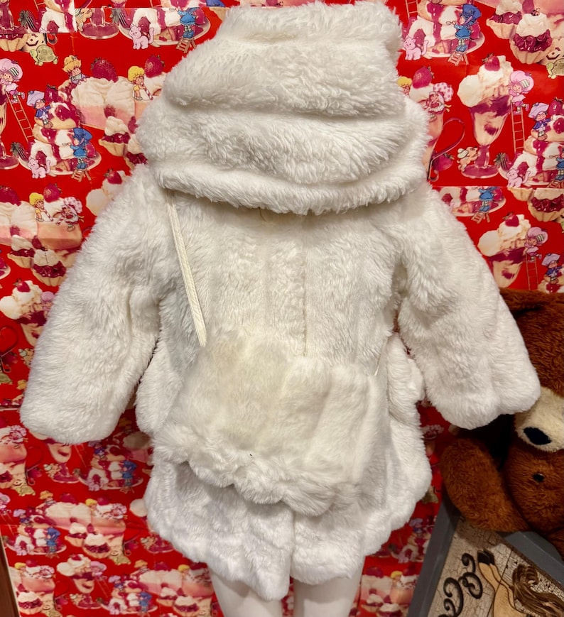 May include: A fluffy white hooded jacket and skirt set. The jacket has long sleeves, a front pocket, and a drawstring. The skirt has a scalloped hem. The outfit is made of a soft, plush material, perfect for a child's winter wardrobe.